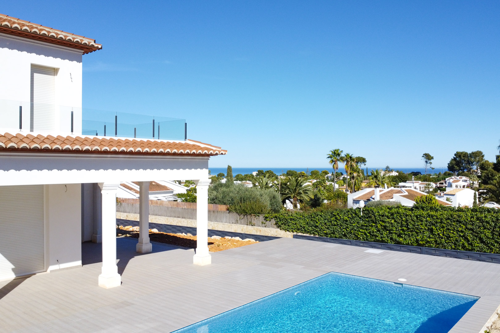 Exterior view featuring a porcelain tiled terrace, blue mosaic pool, and a shaded porch with white columns. The background offers panoramic views of the Mediterranean Sea.