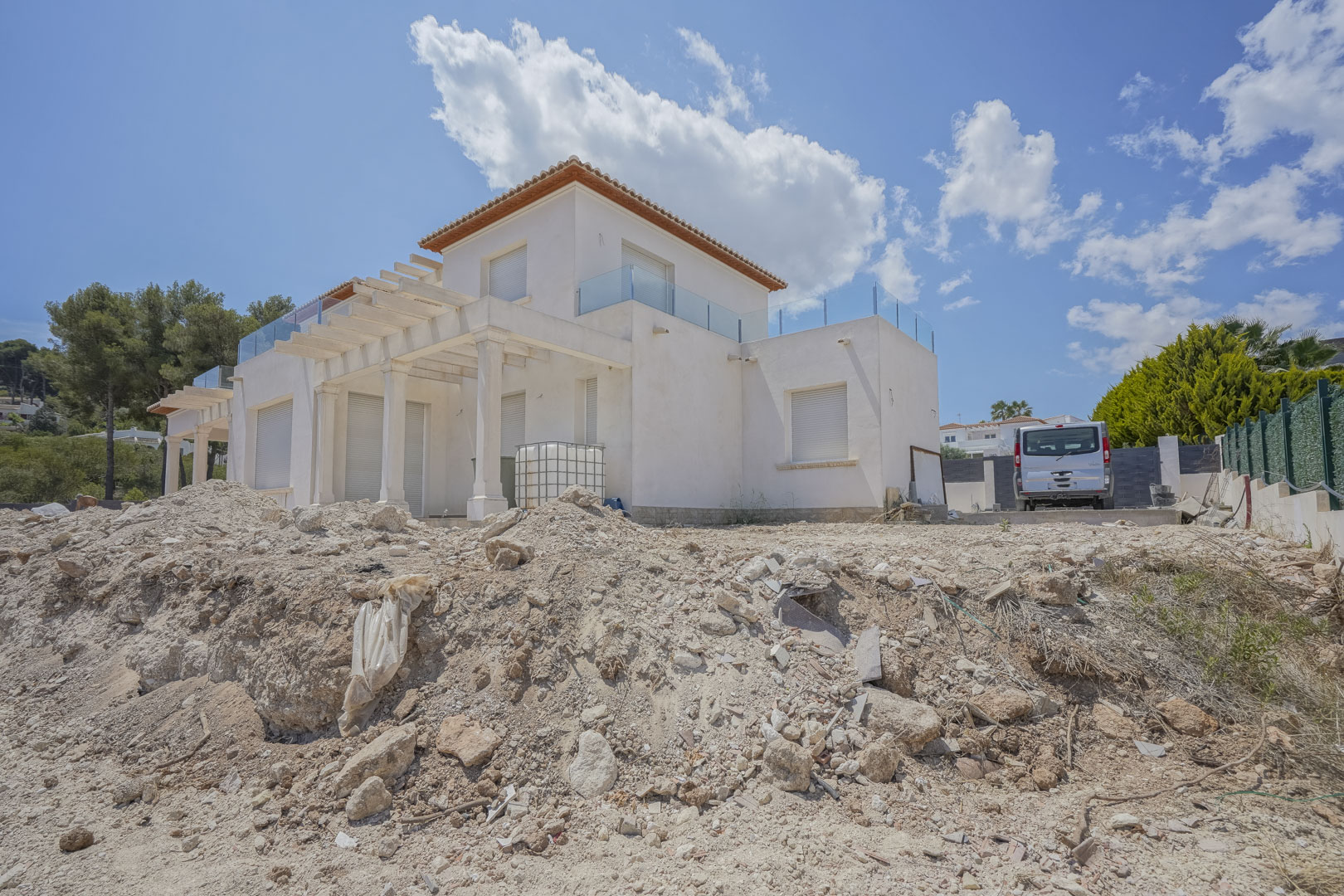 Exterior view of the villa in the final construction stage, featuring white facades, columned pergolas, and upper terraces with modern glass balustrades.