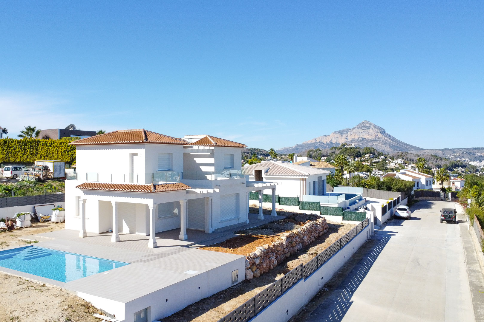Exterior view of the white villa featuring ceramic tile roofing, an integrated pool, and natural stone walls, with the Montgó mountain in the distance.