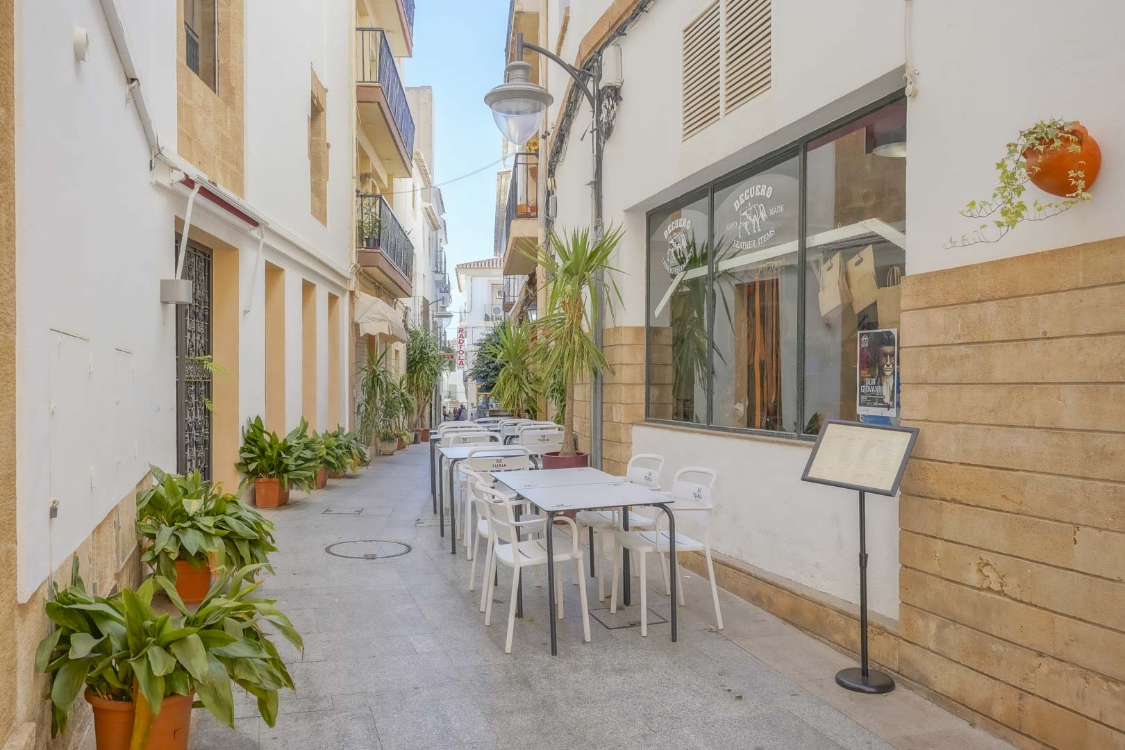 Traditional pedestrian street in the old town featuring Tosca stone walls, potted greenery, and outdoor café seating near local craft shops.