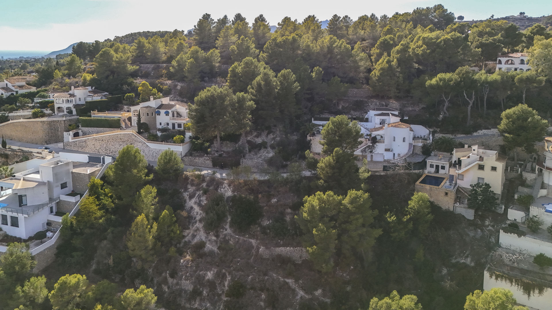 Aerial view of residential plots in Benissa, integrated into a lush pine forest with traditional stone walls and distant Mediterranean Sea views.