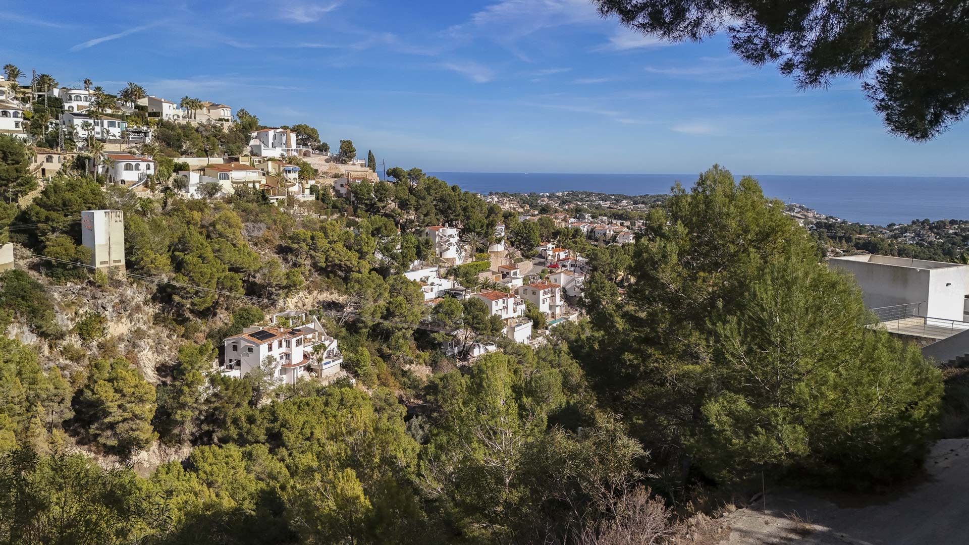 Panoramic view from the elevated plot towards the Mediterranean Sea, featuring white villas nestled in a pine-covered hillside under a clear blue sky.