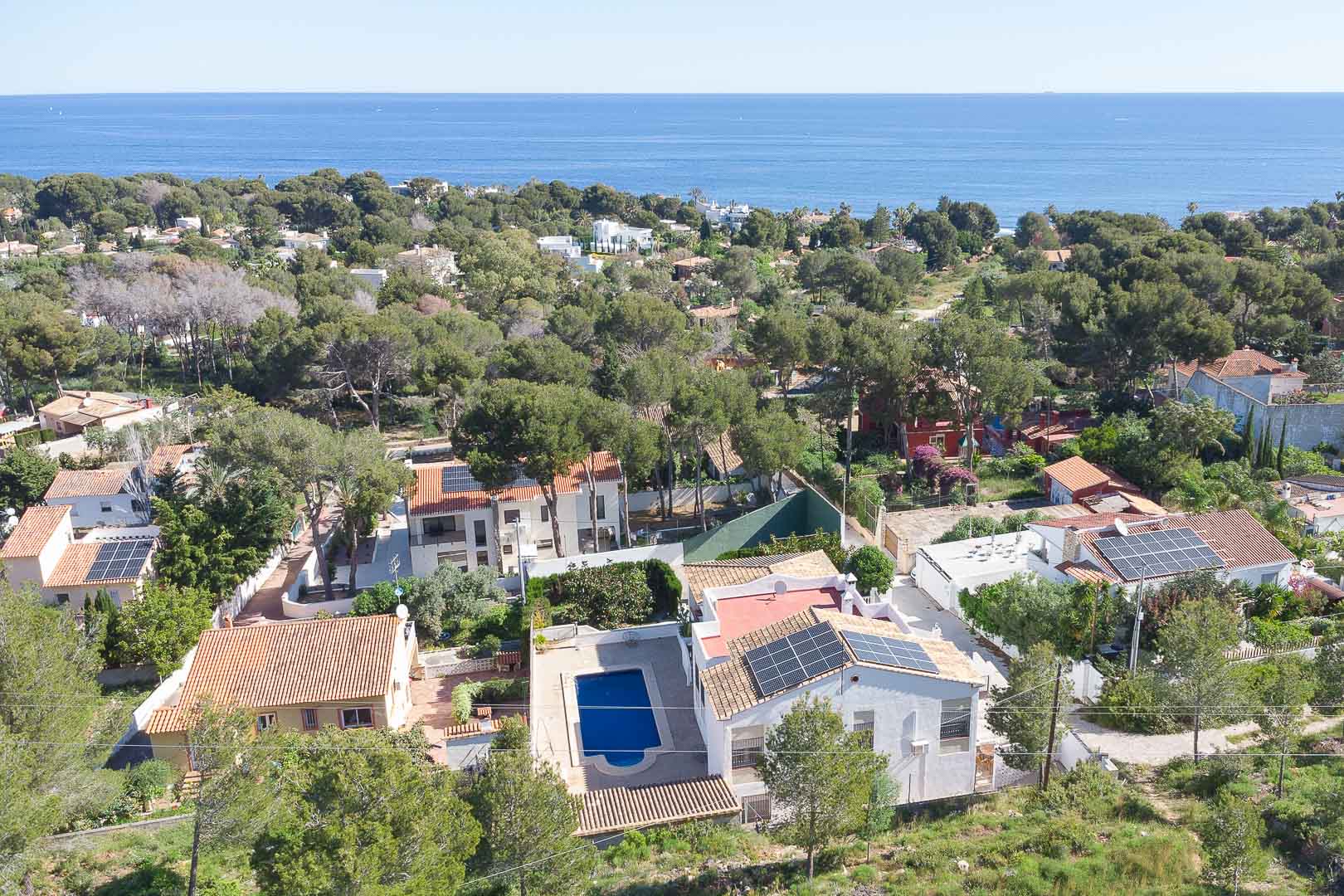 Aerial view of the villa featuring a private pool and rooftop solar panels, set within a lush pine forest in Las Rotas with Mediterranean Sea views.
