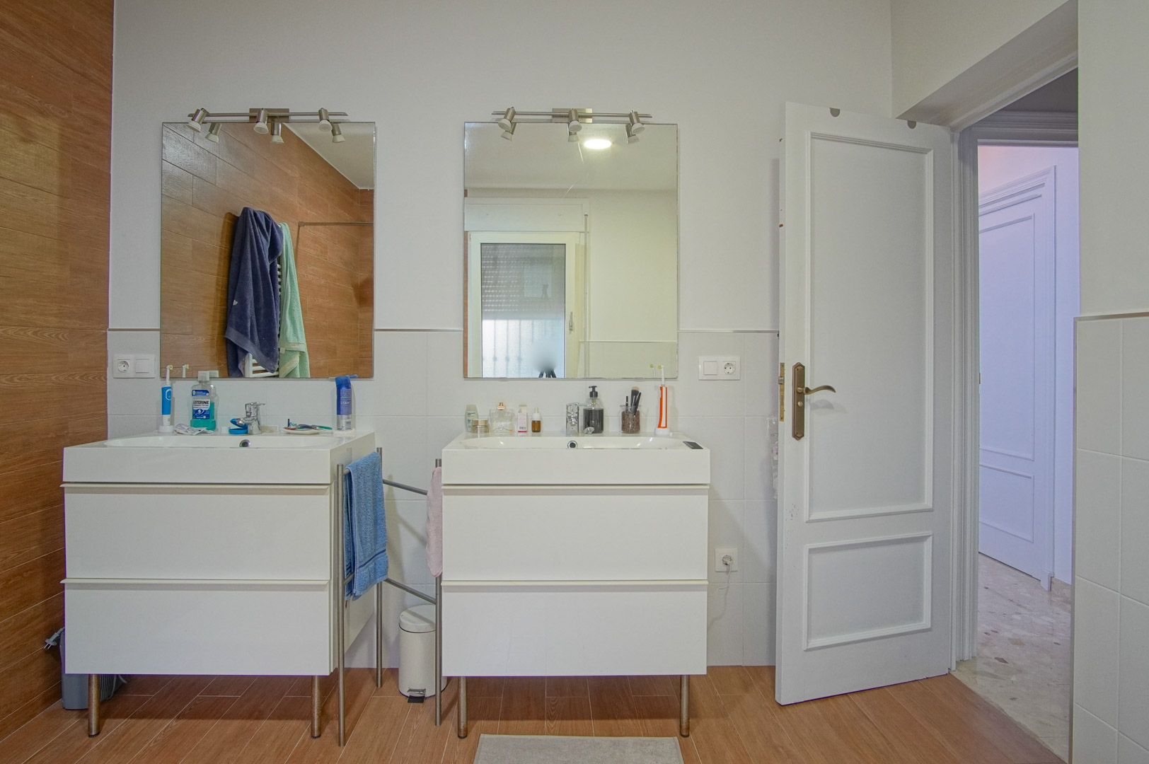Bathroom featuring a double vanity with separate white cabinets, individual mirrors with spotlights, and a combination of white and wood-effect wall tiling.