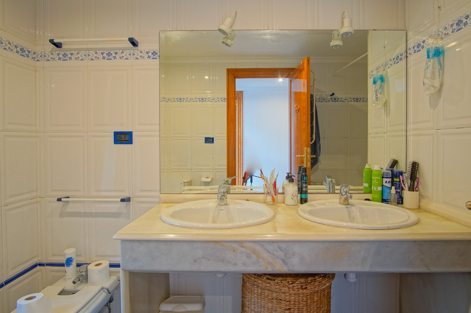 Bathroom featuring a marble double vanity, white tiled walls with a blue floral border, and a large mirror with overhead spotlights.