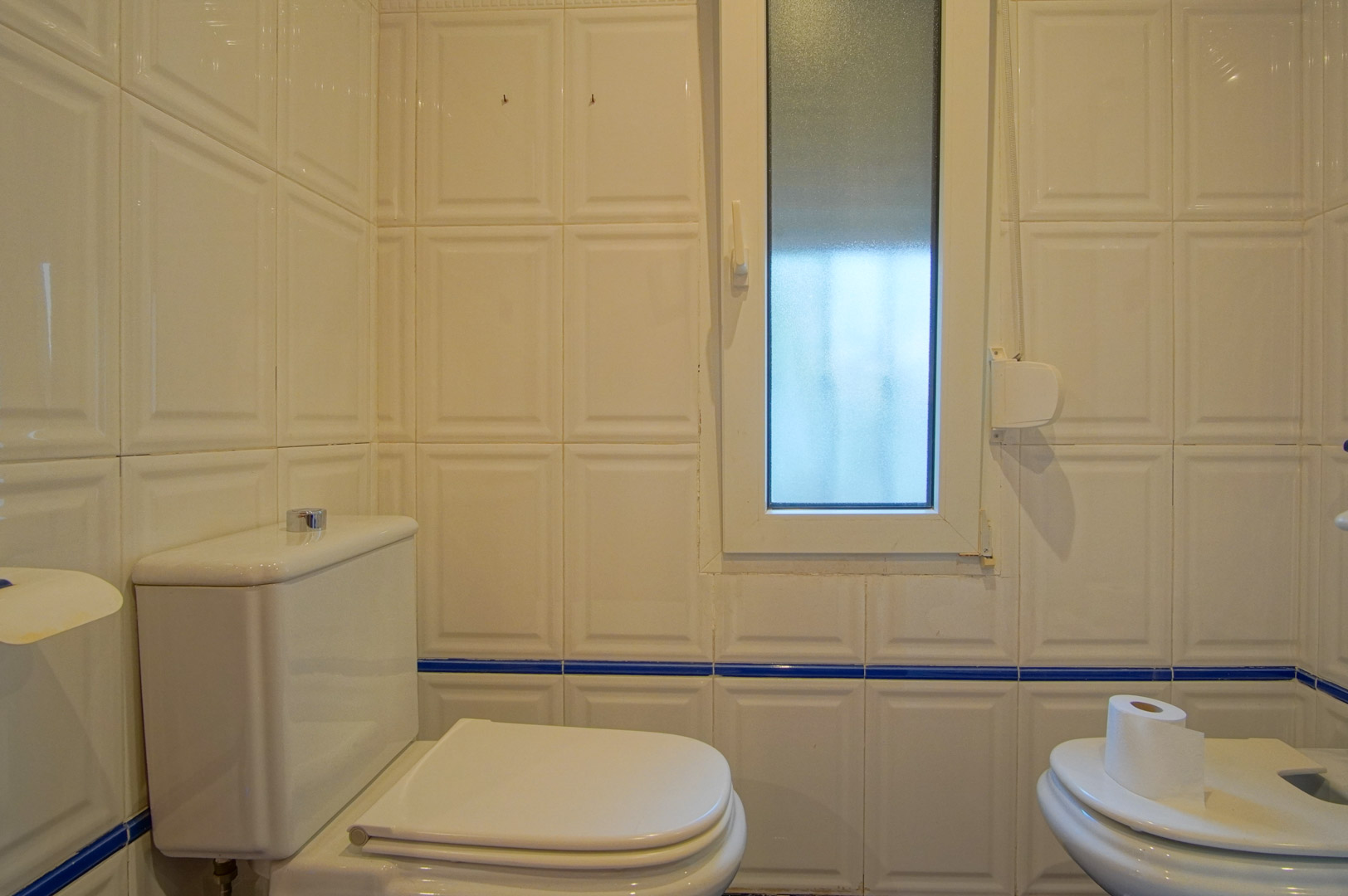 Bathroom featuring white relief wall tiles with a blue border. It includes a toilet, bidet, and a frosted glass window for privacy and natural light.