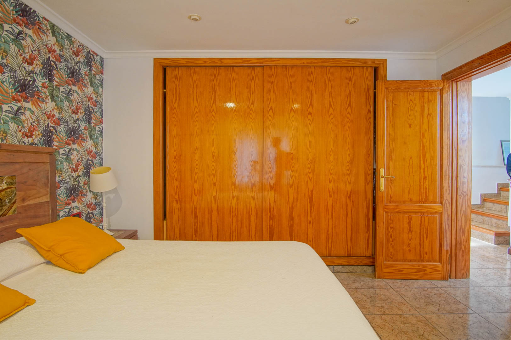 Bedroom featuring a large built-in wooden wardrobe, tropical floral accent wall, and tiled flooring. The open door reveals a hallway leading to the staircase.