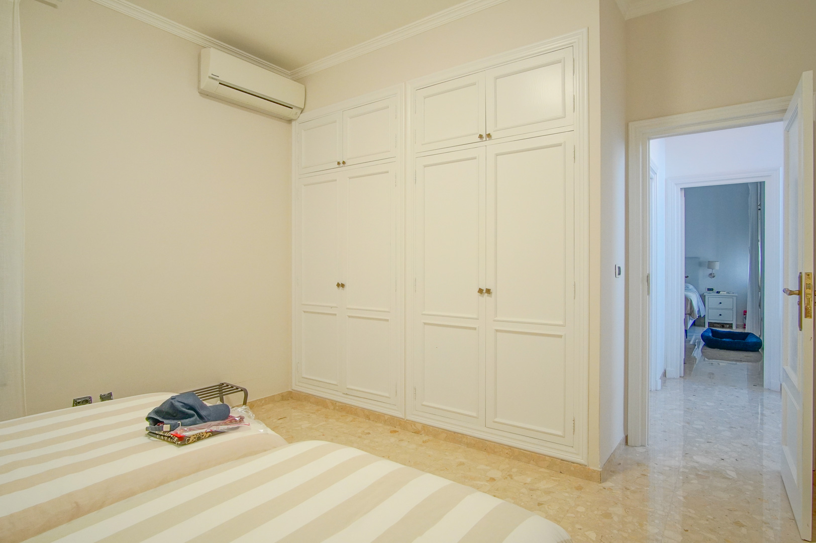 Bedroom featuring large white built-in wardrobes, polished marble flooring, and wall-mounted air conditioning, connected to the main hallway.