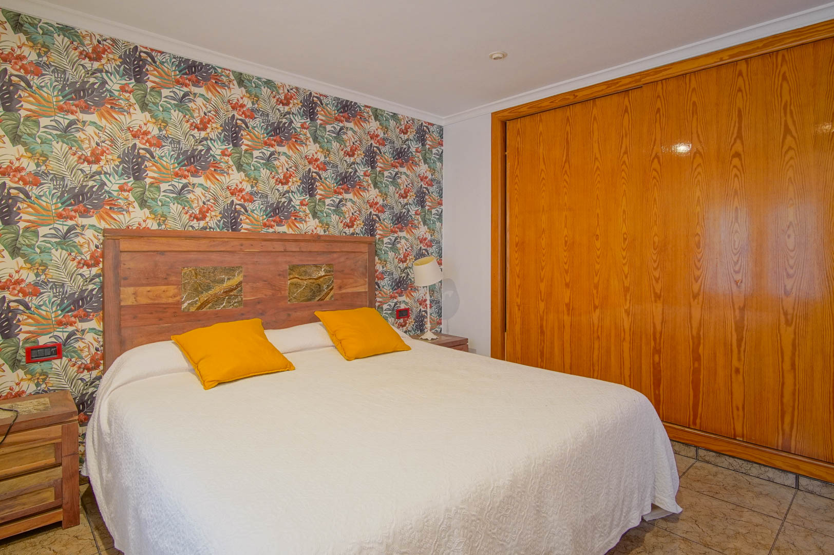 Bedroom featuring tropical-themed wallpaper, a rustic wooden headboard with stone details, and a large built-in wooden wardrobe with sliding doors.