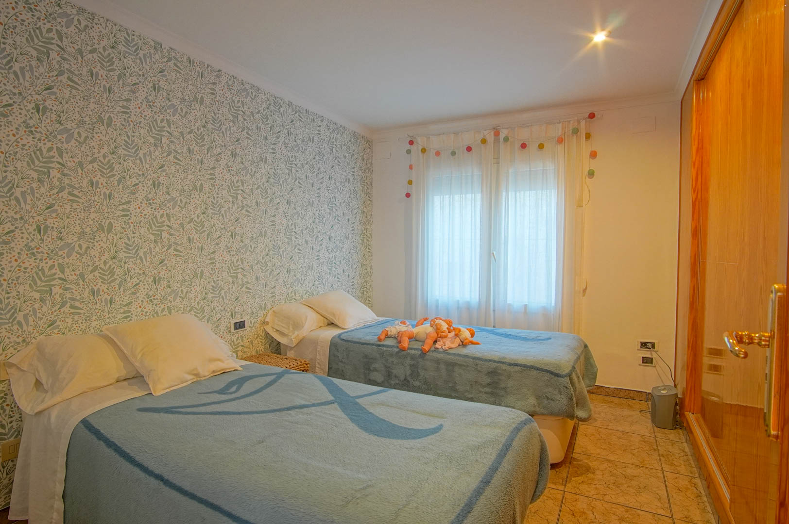 Bedroom with twin beds, botanical accent wallpaper, built-in wooden sliding wardrobes, and natural light filtered through white curtains.