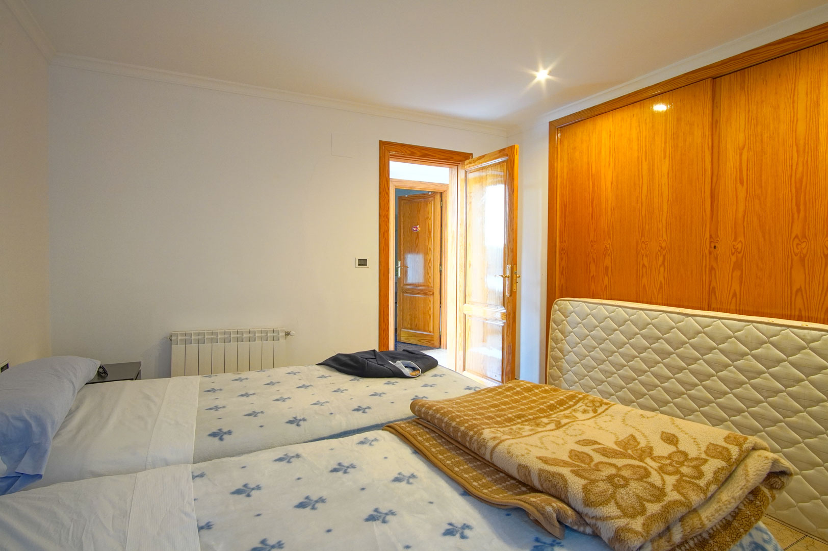 Bright bedroom featuring large built-in wooden wardrobes, twin beds, and a wall-mounted radiator. Matching wooden doors and recessed lighting enhance the warm interior.