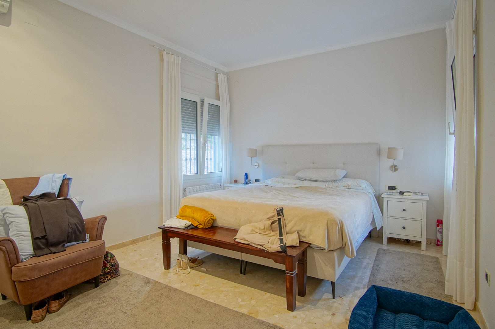 Bright bedroom featuring marble flooring, a double bed with an upholstered headboard, and a wooden footbench. Two windows with shutters provide ample natural light.