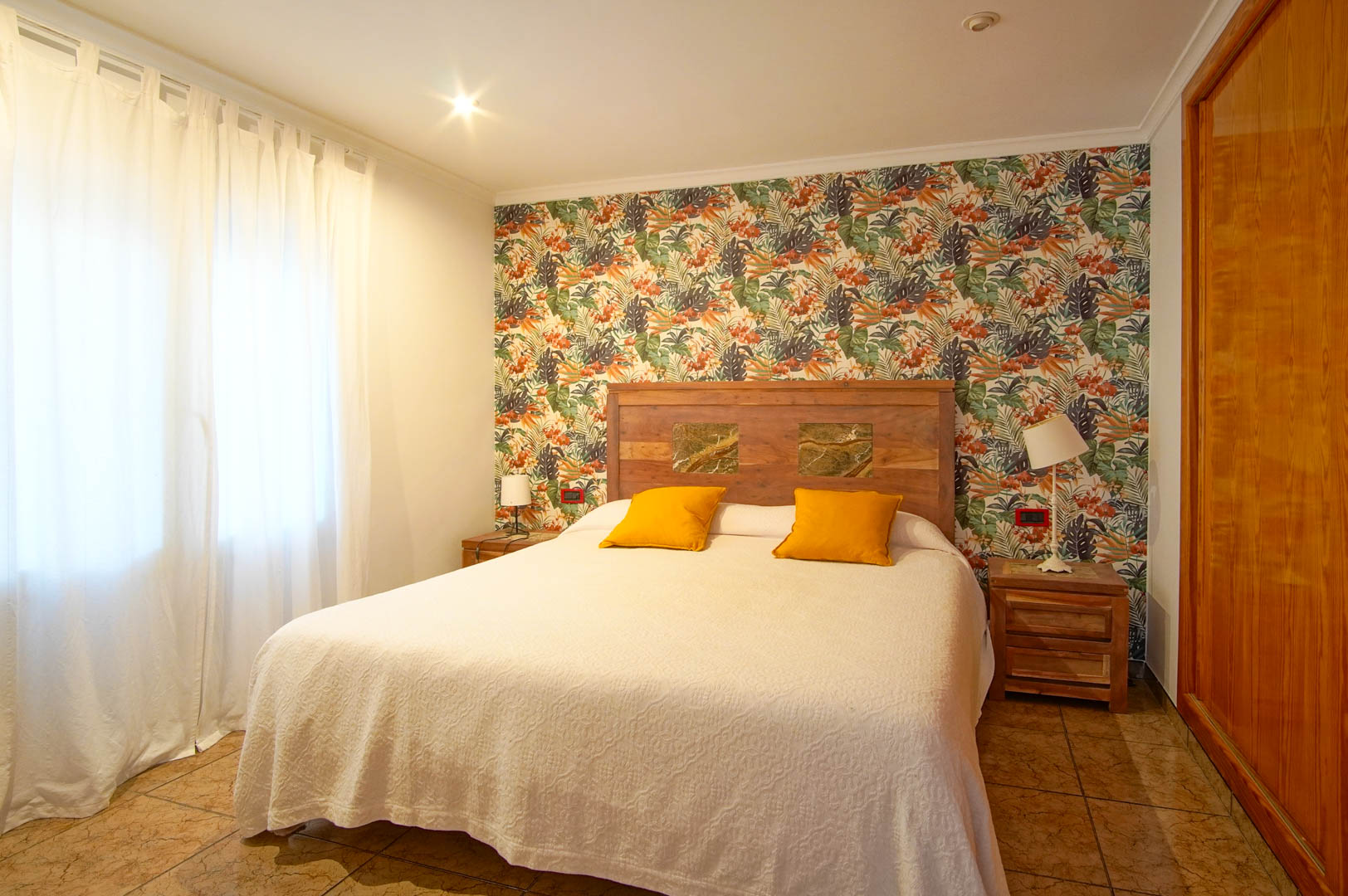 Bright bedroom featuring tropical patterned wallpaper, a wooden headboard with stone inlays, and integrated wooden wardrobes.