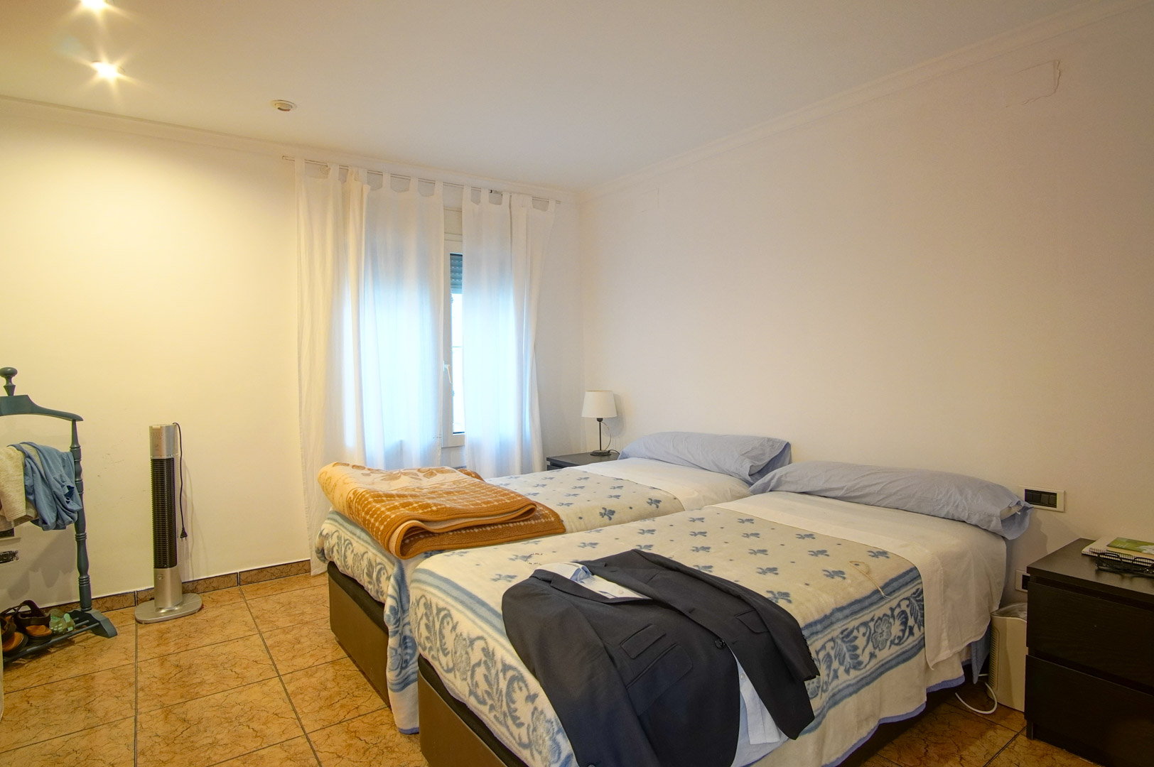 Bright bedroom featuring twin beds, ceramic tile flooring, and white walls. The space includes recessed ceiling lighting and a window providing soft natural light.