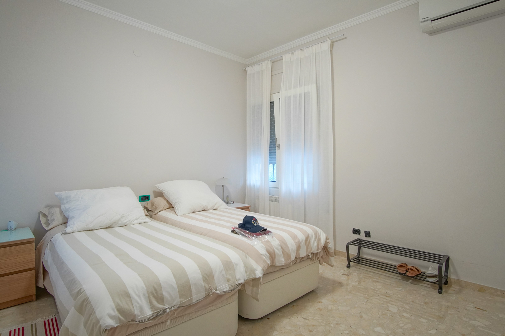Bright bedroom featuring twin beds with striped linens, marble flooring, split-system air conditioning, and a window with translucent white curtains.