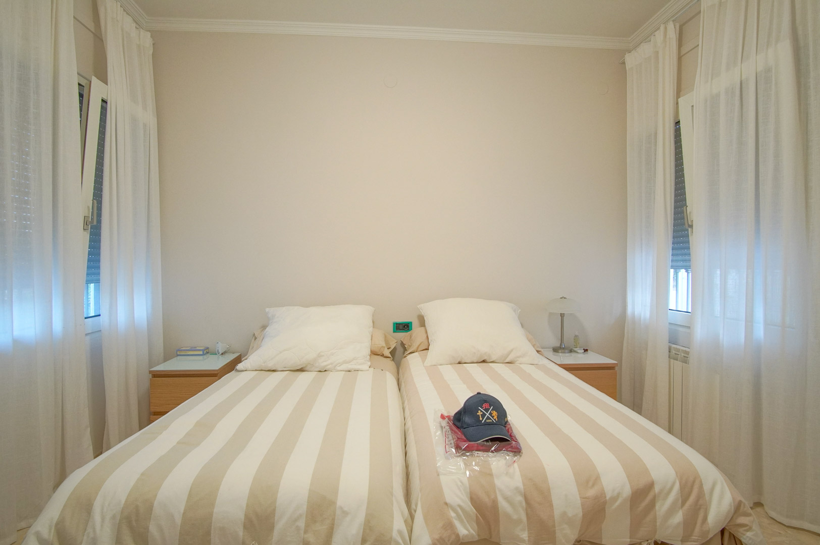 Bright bedroom featuring twin beds with striped linens, wooden nightstands, and dual windows framed by soft white curtains for natural illumination.