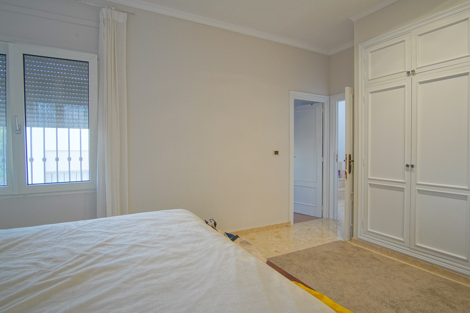 Bright bedroom featuring white built-in wardrobes, marble flooring, and a window with integrated shutters. The neutral color palette creates a serene atmosphere.