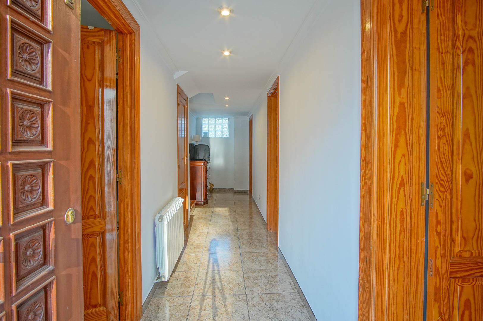 Bright hallway featuring ceramic tile flooring, carved wooden doors, and recessed ceiling lighting. A white radiator and a glass block window complete the space.
