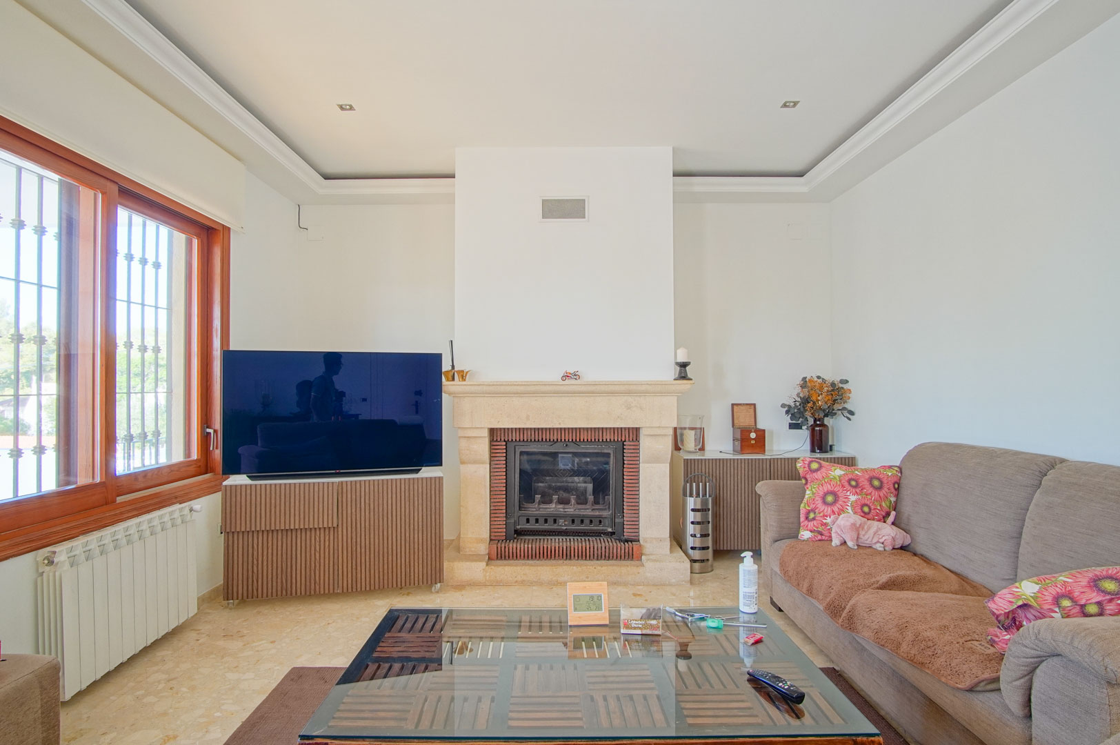 Bright living room featuring a stone fireplace, marble flooring, and tray ceilings with recessed lighting. Large wooden-framed windows provide abundant natural light.