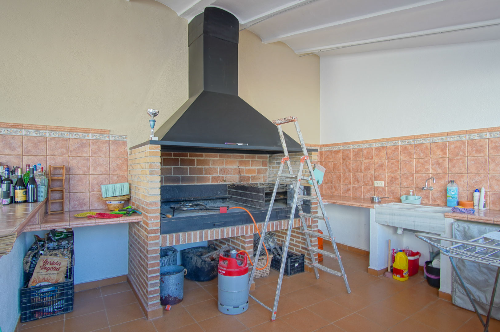 Built-in brick barbecue area featuring a large black metal hood, stone sink, and terracotta-tiled worktops designed for outdoor catering.