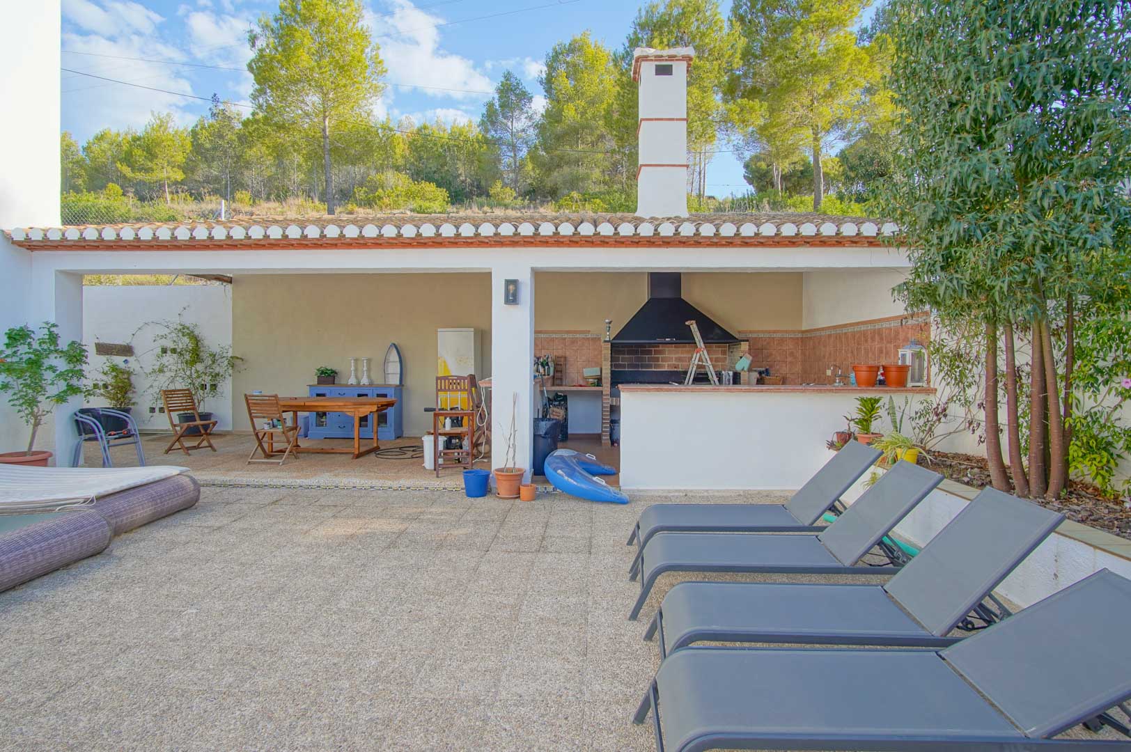 Covered outdoor kitchen and BBQ area with a dining zone and stone terrace with sun loungers, set against the pine forest of the Montgó Natural Park.