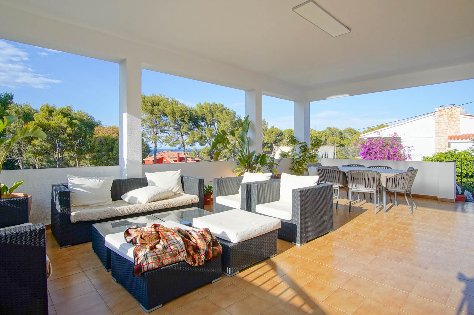 Covered terrace featuring a wicker lounge set, outdoor dining area, and tiled flooring, overlooking the Mediterranean pine forest and the sea.