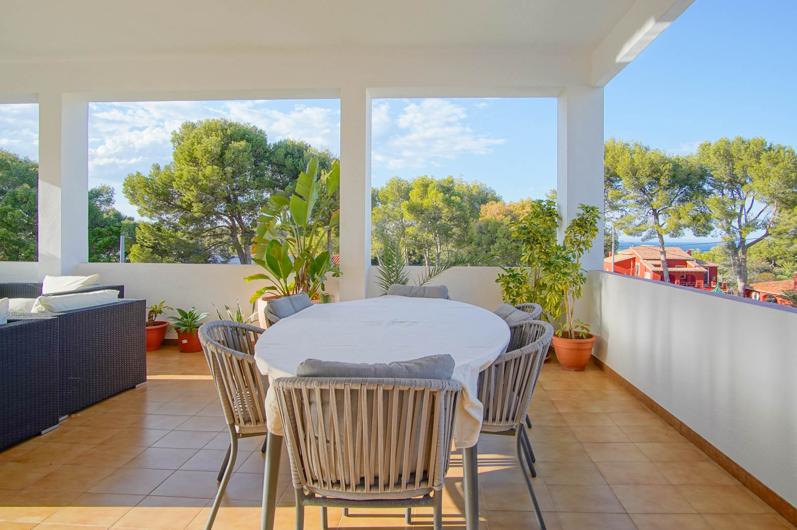 Covered terrace with an outdoor dining set and wicker lounge, providing clear views of the lush Mediterranean pine forest and the distant sea horizon.