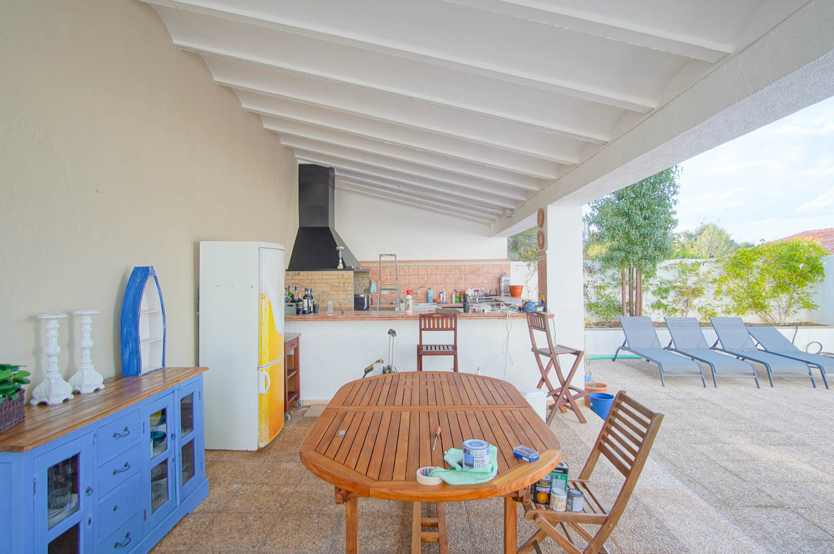 Covered terrace with an outdoor kitchen, built-in BBQ, and white beamed ceiling. Features a wooden dining set overlooking the poolside lounging area and garden.
