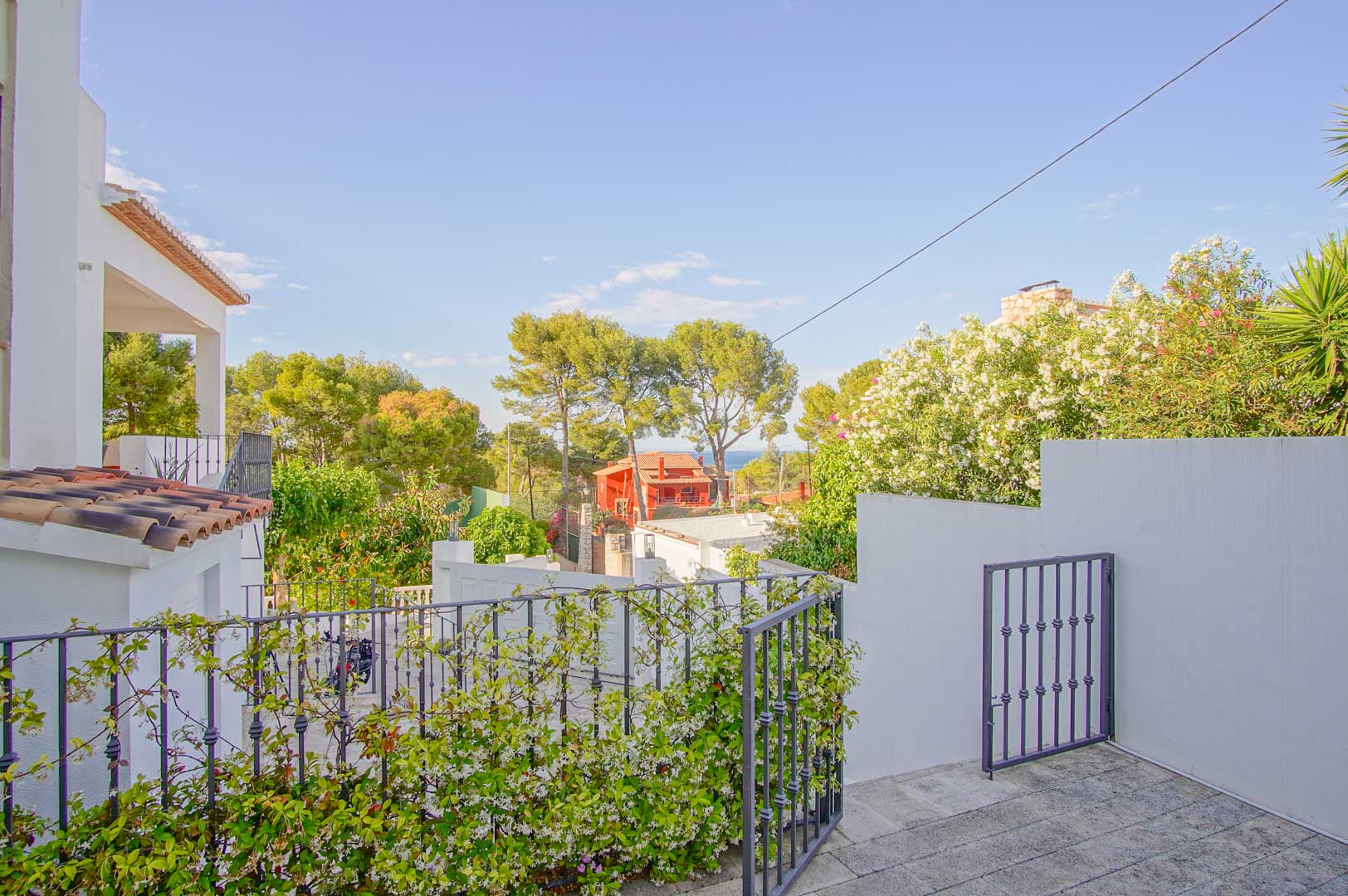 Elevated terrace view featuring stone paving and wrought iron railings with climbing jasmine, offering a glimpse of the Mediterranean Sea through the lush pine trees.