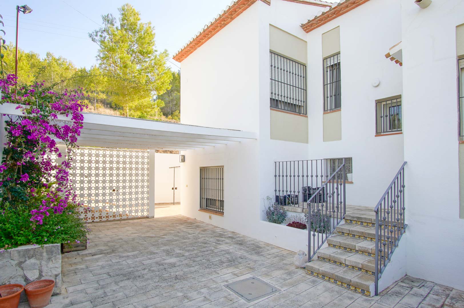 Exterior entrance featuring stone paving, a white pergola-covered parking area, and access stairs with decorative tile risers and wrought iron railings.