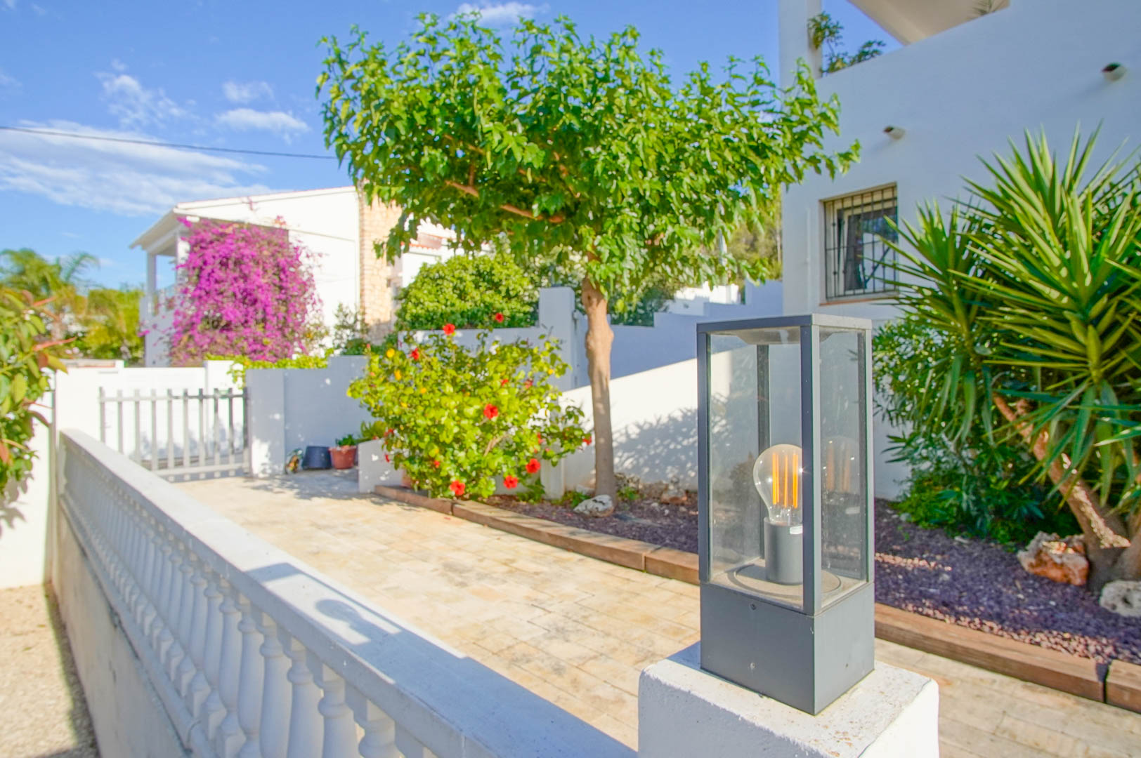 Exterior entrance featuring stone paving, modern glass lantern lighting, and Mediterranean landscaping with hibiscus and vibrant bougainvillea against white walls.