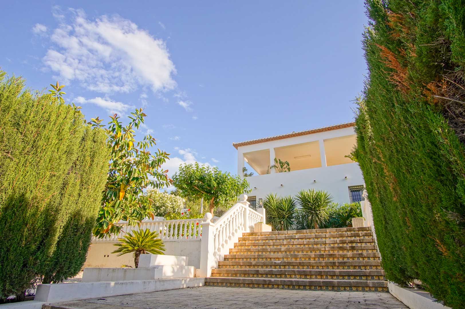 Exterior stone staircase with decorative tile risers and white balustrades, framed by tall cypress hedges leading toward the white villa facade.