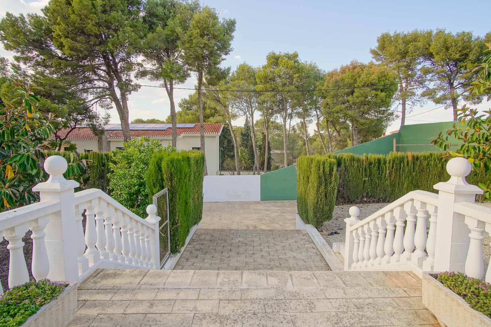 Exterior stone staircase with white balustrades leading to the driveway, framed by mature pine trees, manicured hedges, and the private fronton court.