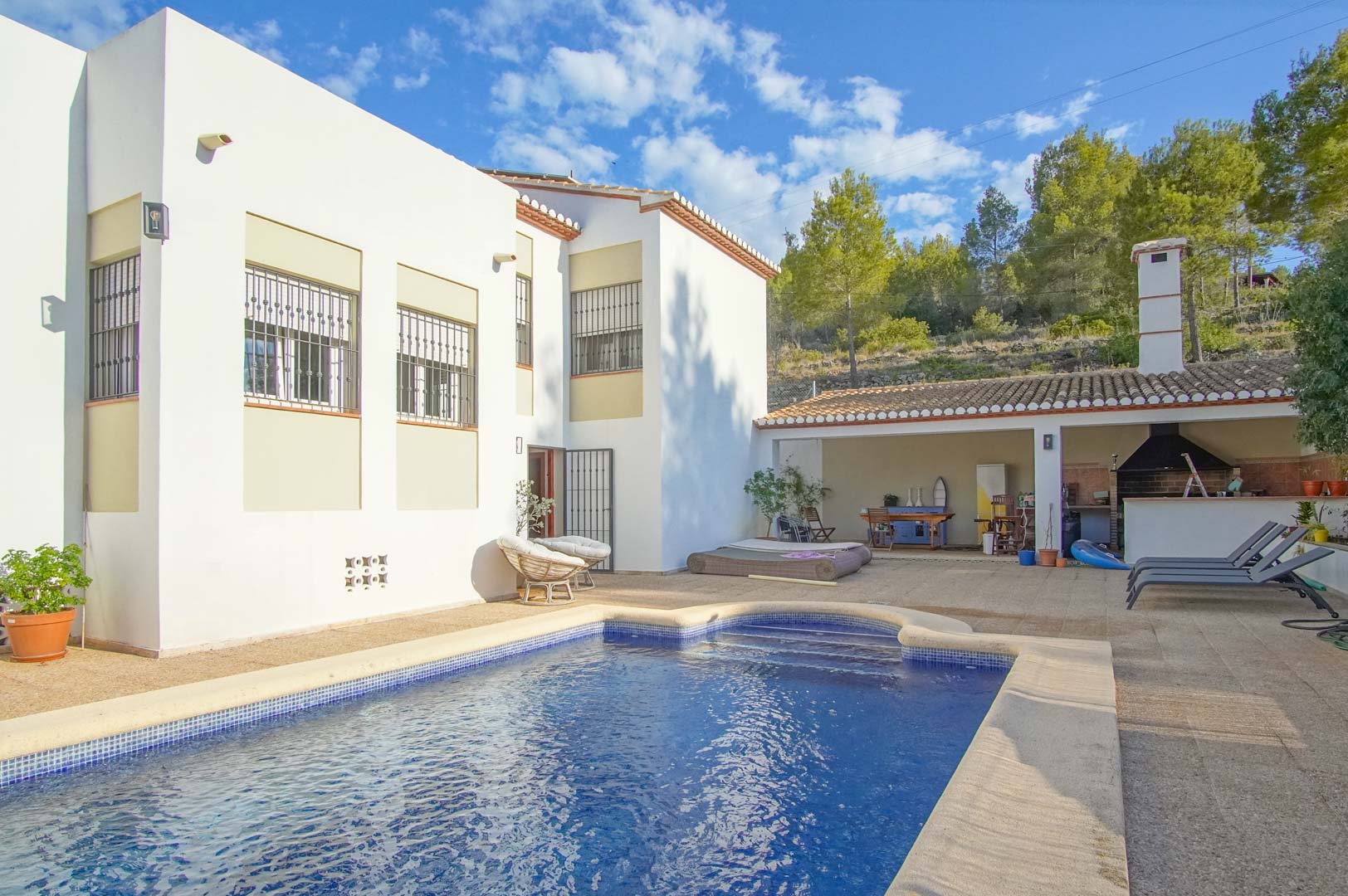 Exterior view featuring a private swimming pool, spacious sun terrace, and a covered outdoor kitchen with BBQ area, set against a pine forest backdrop.