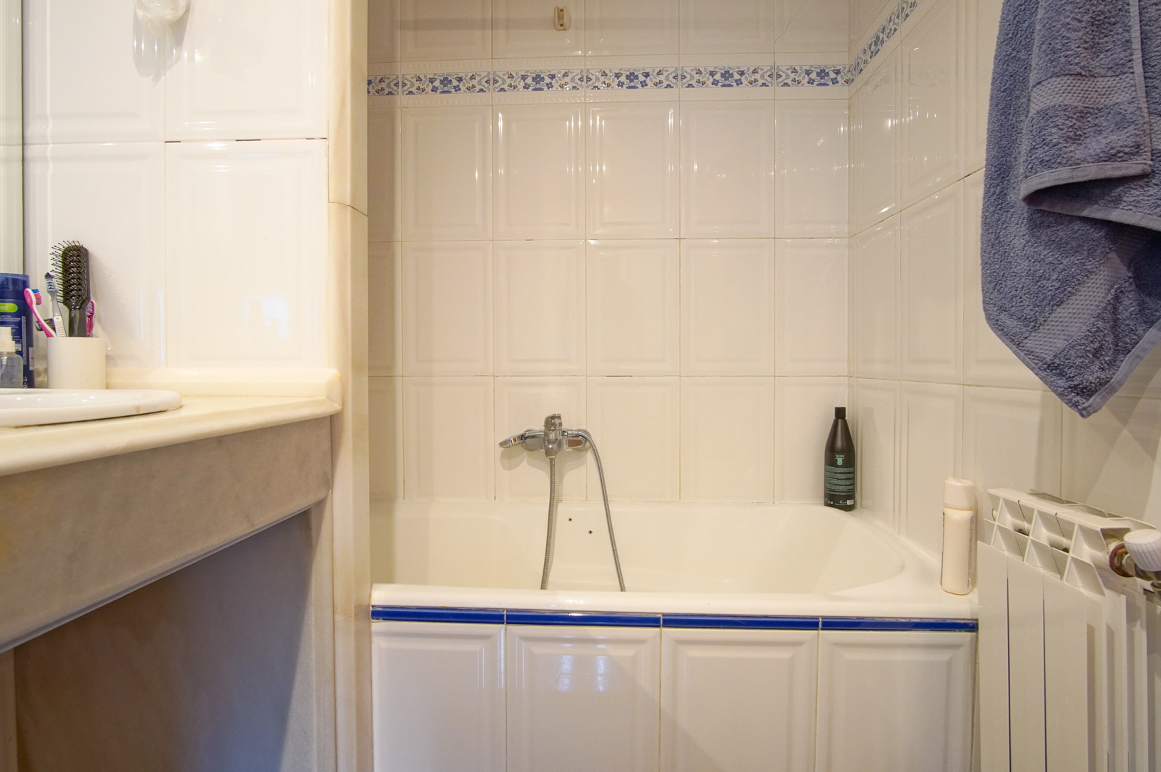 Functional bathroom with a built-in bathtub, white ceramic wall tiles featuring blue floral accents, and a marble vanity top. Includes a radiator and chrome fixtures.