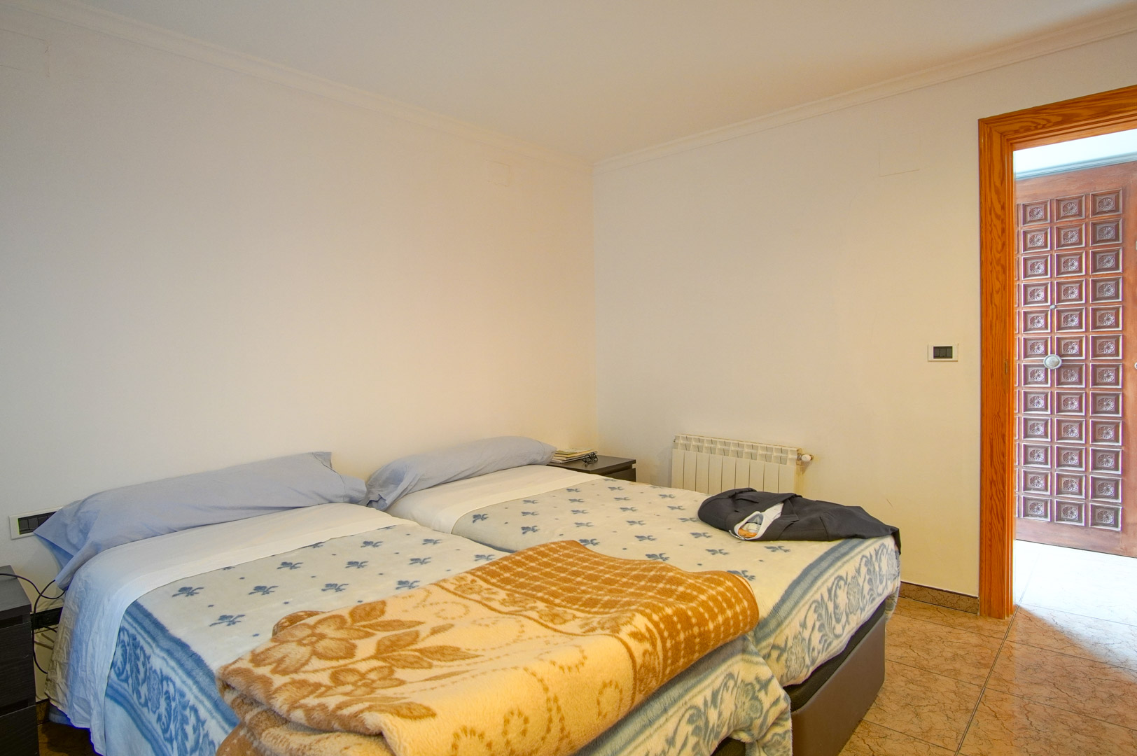 Functional bedroom with twin beds, white walls, and ceramic tile flooring. It features a wall radiator and a distinctive carved wooden panel door.