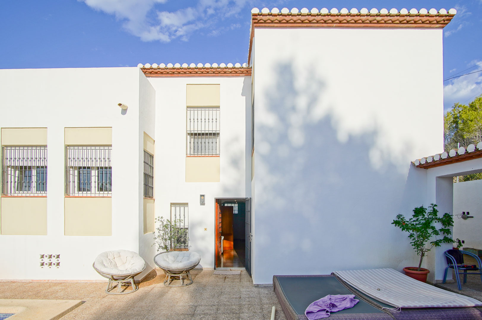 Mediterranean-style white facade featuring barred windows, a stone-paved terrace with outdoor seating, and a direct entrance under a clear blue sky.