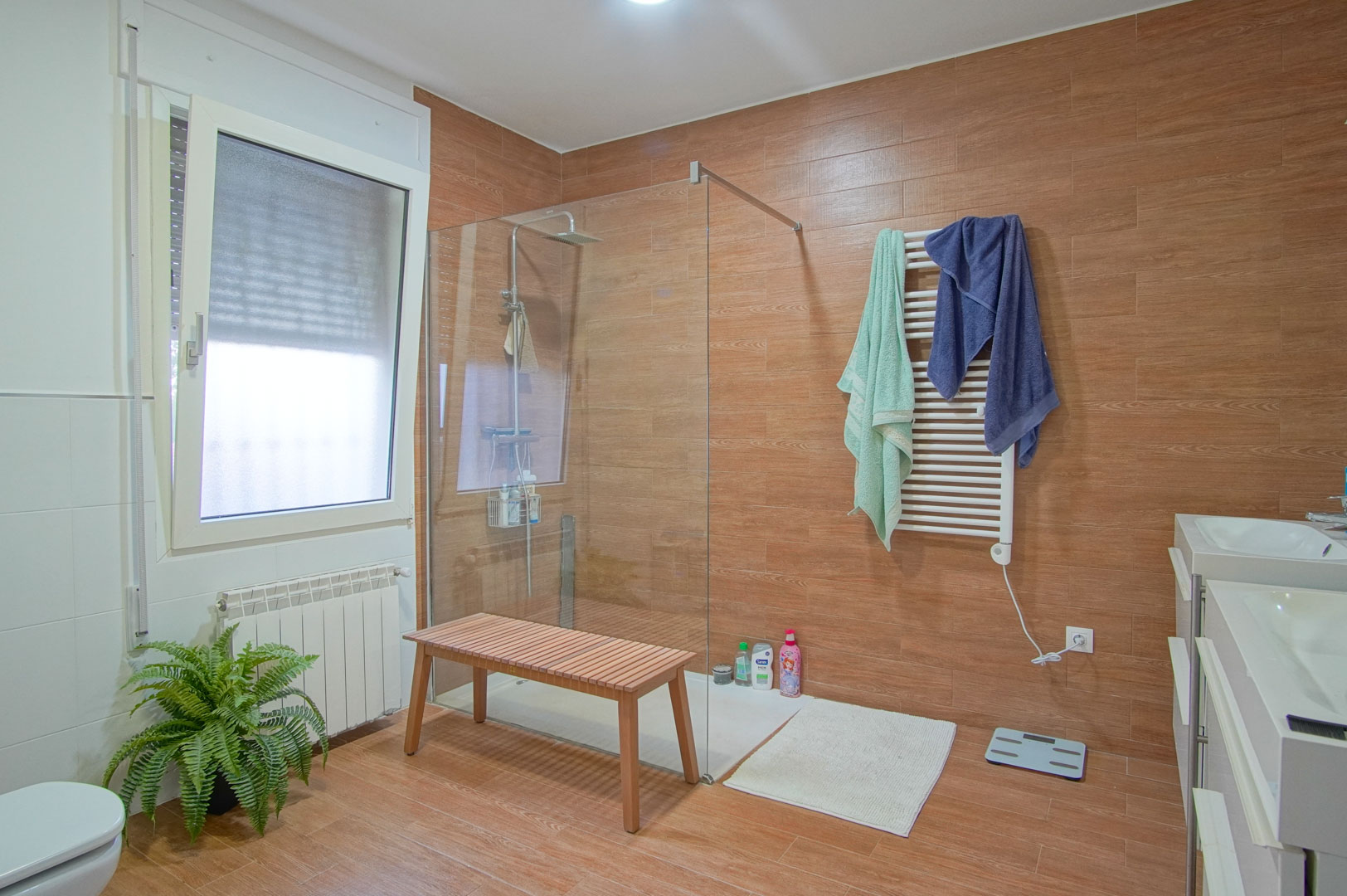 Modern bathroom featuring wood-effect tiling, a walk-in glass shower, double vanity sinks, and a white heated towel rail for added comfort.