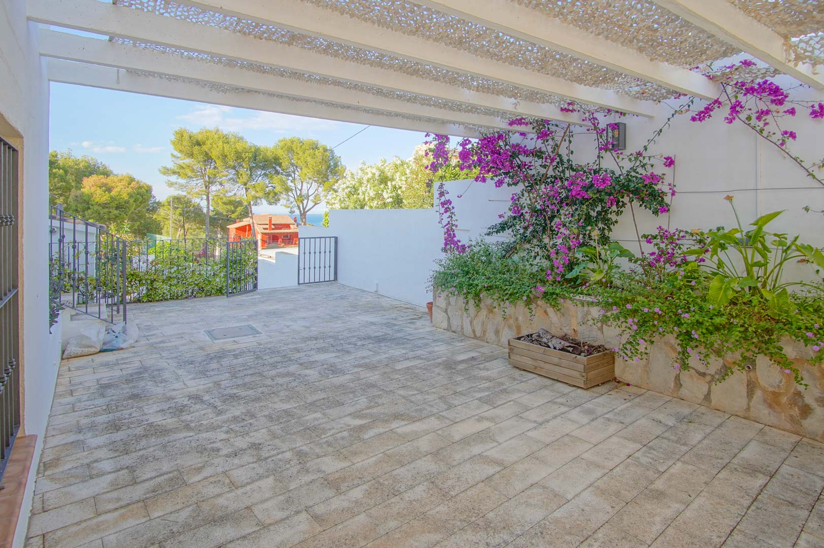 Paved outdoor terrace featuring a white pergola with shade netting, stone-clad planters with bougainvillea, and distant sea views framed by Mediterranean pine trees.