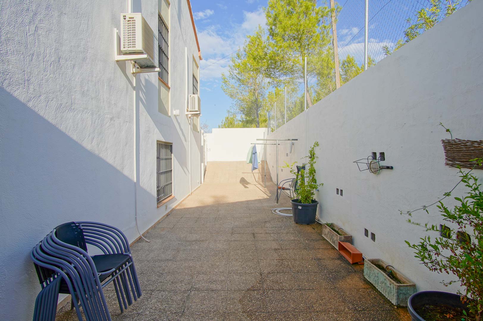 Paved side patio featuring white exterior walls, wall-mounted air conditioning units, and a high perimeter fence bordered by Mediterranean pine trees.
