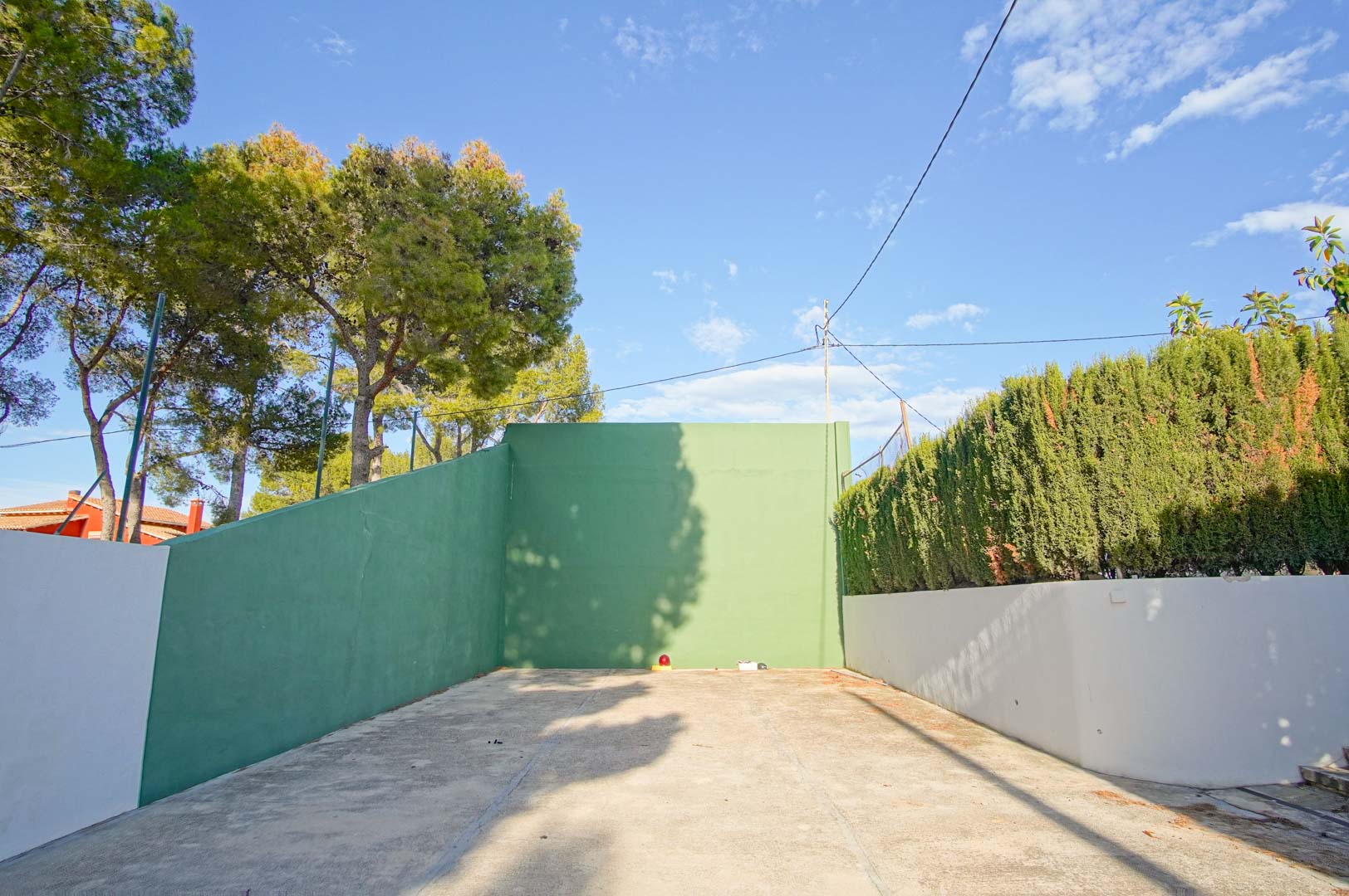 Private fronton court featuring green walls and a concrete surface, bordered by mature pine trees and high hedges for seclusion.