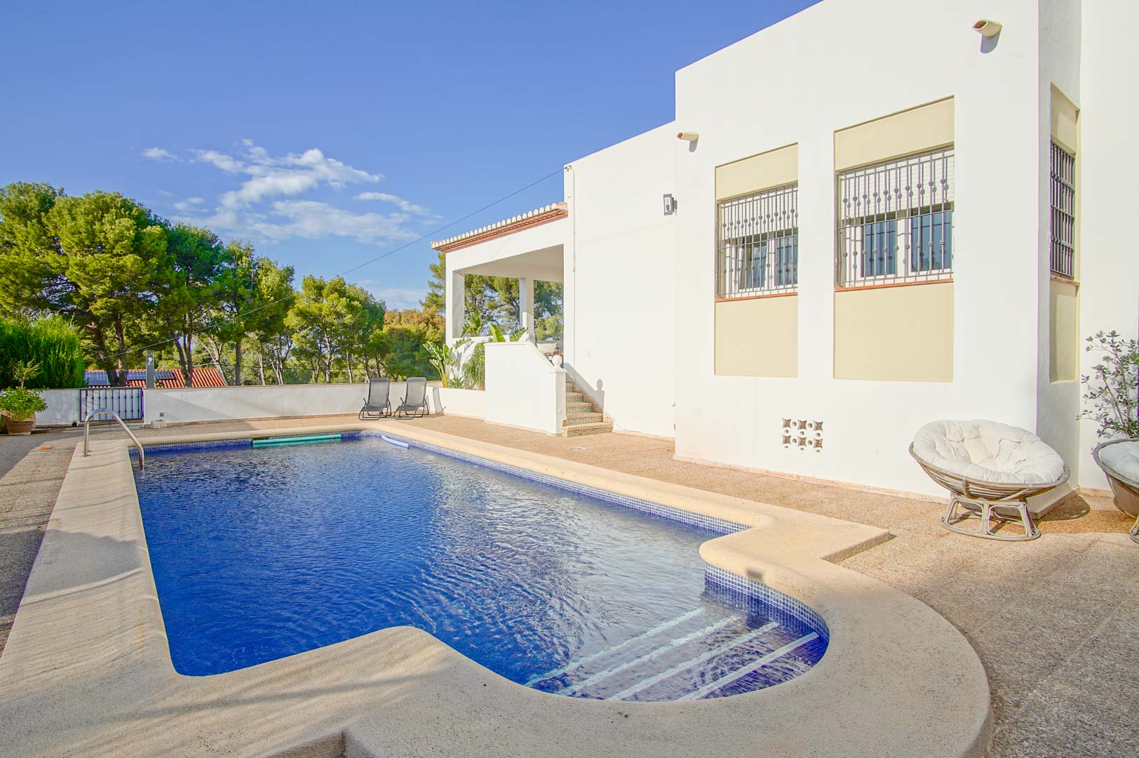 Private pool with blue mosaic tiling and a stone terrace, adjacent to the white villa facade and surrounded by lush Mediterranean pine trees.