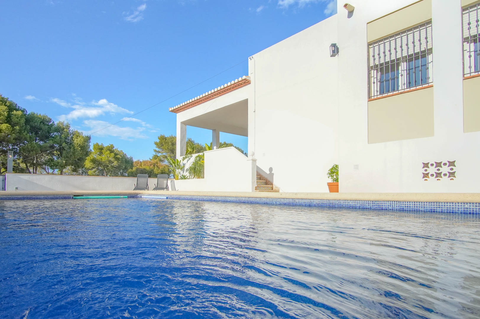 Private swimming pool with blue mosaic tiling adjacent to the white villa facade, featuring a covered terrace, tiled stairs, and a backdrop of Mediterranean pine trees.