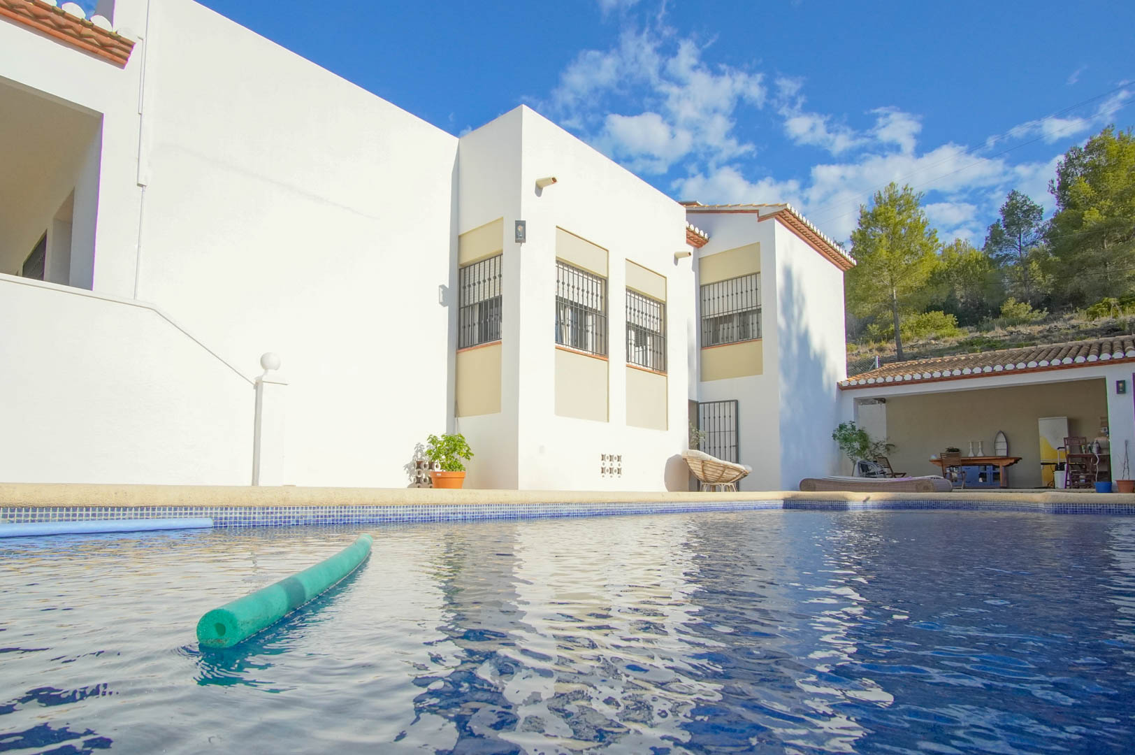 Private swimming pool with blue mosaic tiling and a spacious sun deck adjacent to the white villa facade and a covered outdoor lounge area.