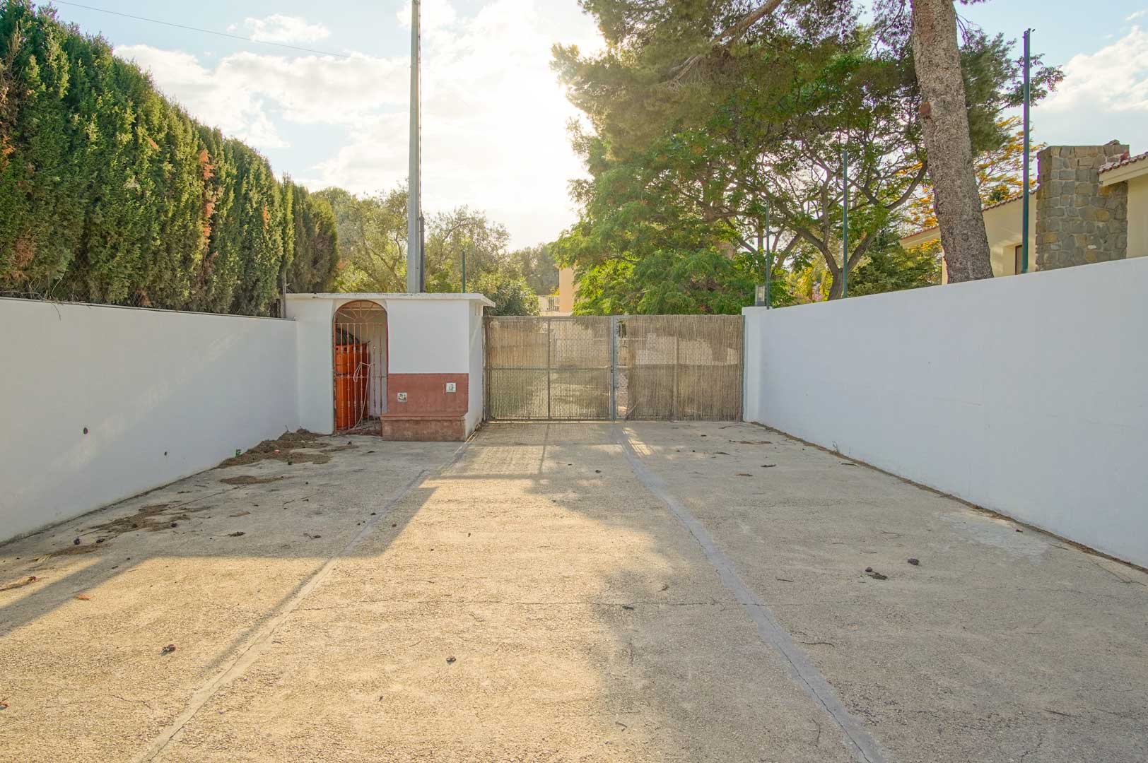 Property entrance featuring a paved driveway, white perimeter walls, and a gated access point framed by mature pine trees and tall cypress hedges.