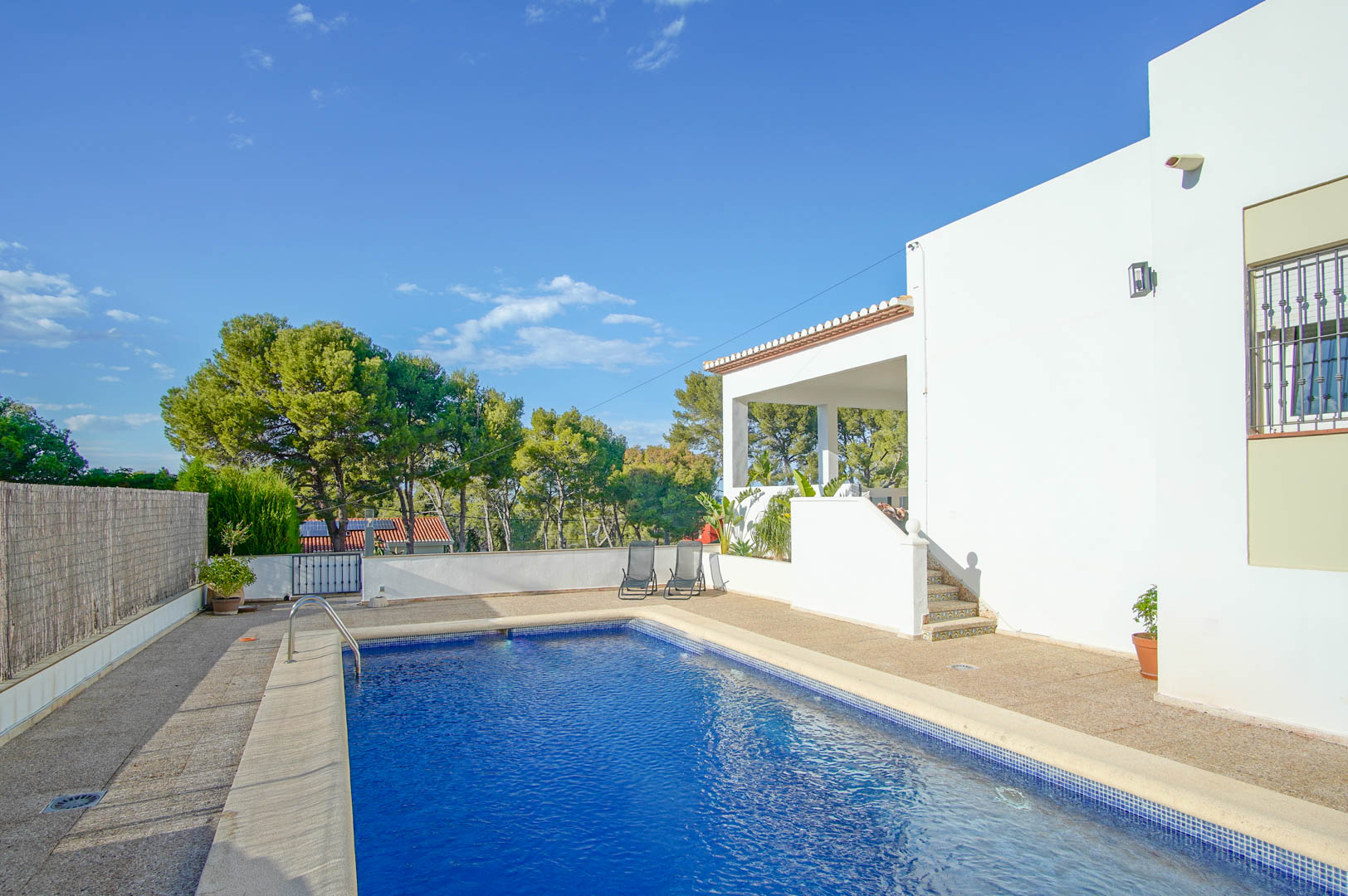 Rectangular private pool with blue mosaic tiling, featuring a spacious stone-paved sun terrace and stairs leading to the elevated porch of the white villa.