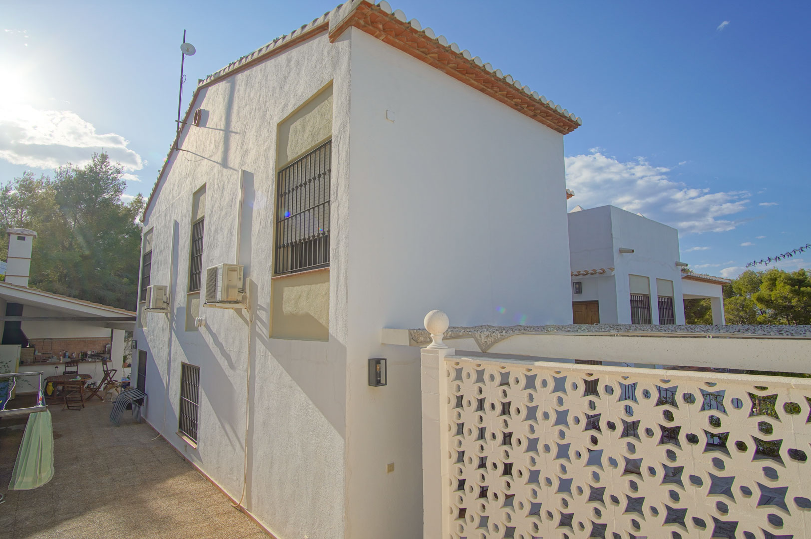 Side facade of the villa featuring white walls, barred windows, and a paved walkway leading toward an outdoor kitchen and barbecue area.