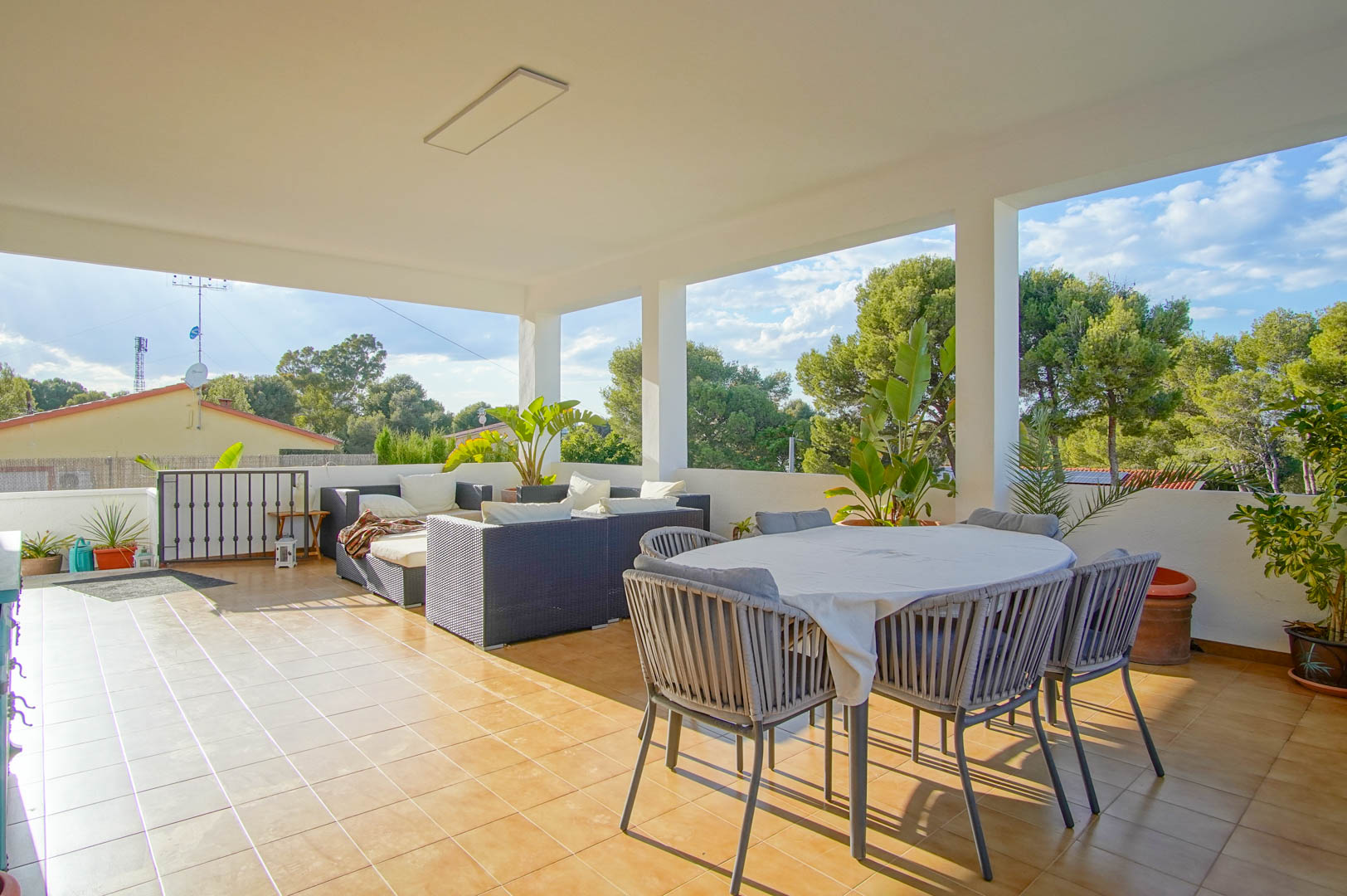 Spacious covered terrace featuring a dining area, wicker lounge furniture, and ceramic tiling, offering views of the surrounding Mediterranean greenery.