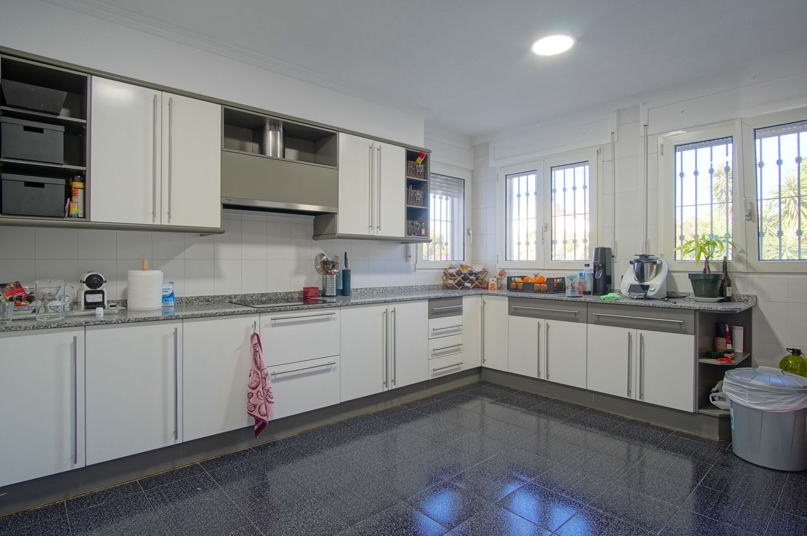 Spacious independent kitchen featuring white and grey cabinetry, granite countertops, and dark tiled flooring. Multiple windows provide ample natural light to the workspace.