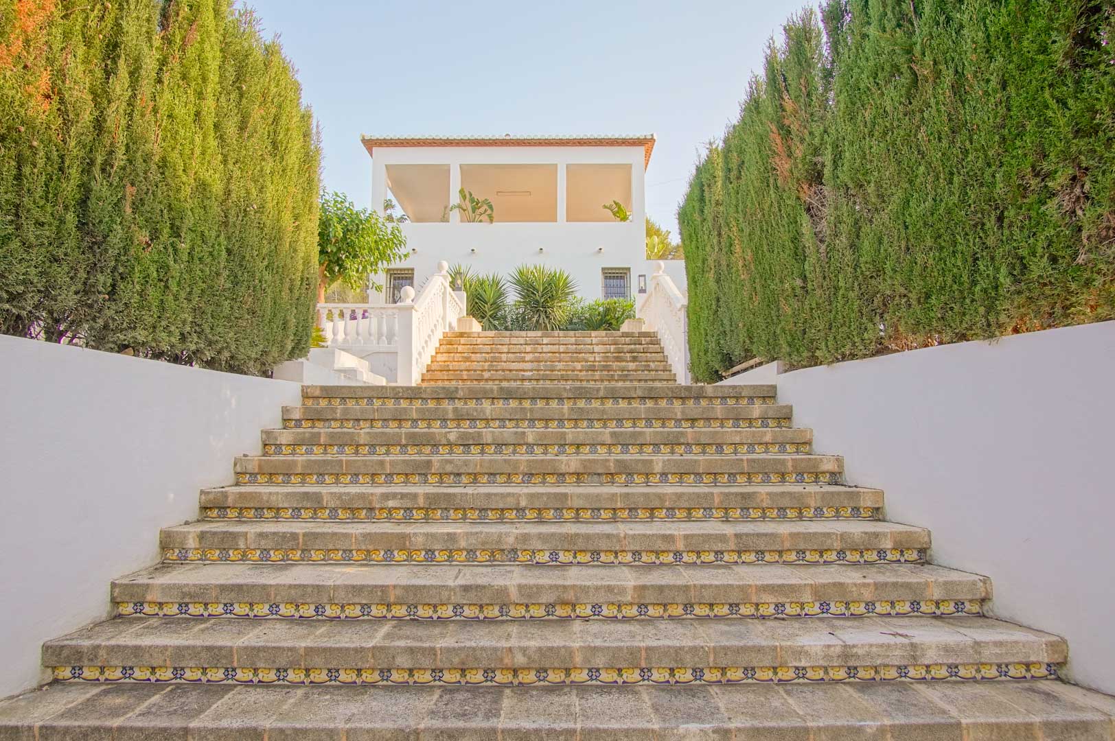 Wide stone staircase featuring decorative tile risers, bordered by tall cypress hedges leading toward the villa's white facade and arched upper terrace.
